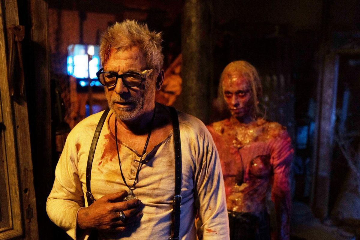 A disheveled man with glasses and blood all over his shirt holds a pendant around his neck with a creepy zombie-like figure in the background.